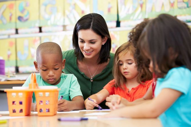 woman watching young children color with crayons in the classroom