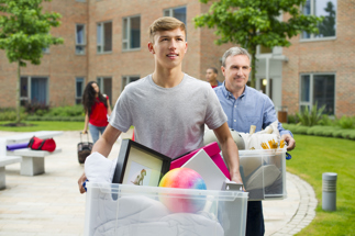Young adult male carrying box of dormitory items with adult male in background